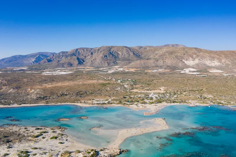 Aerial view of the Elafonisi lagoon: turquoise shallows and a pale sandbar running out towards a low islet, with bare hills behind.