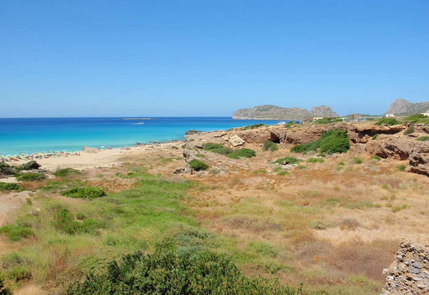 Falassarna seen from the dunes: a long pale beach, turquoise water and a rocky headland offshore.