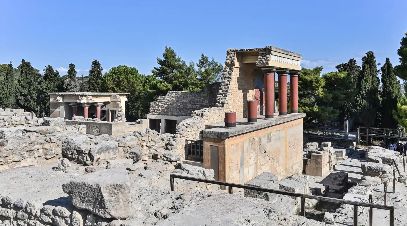 The reconstructed north entrance of the Minoan palace at Knossos: red columns under a stone portico, among excavated foundations and pine trees under a blue sky.