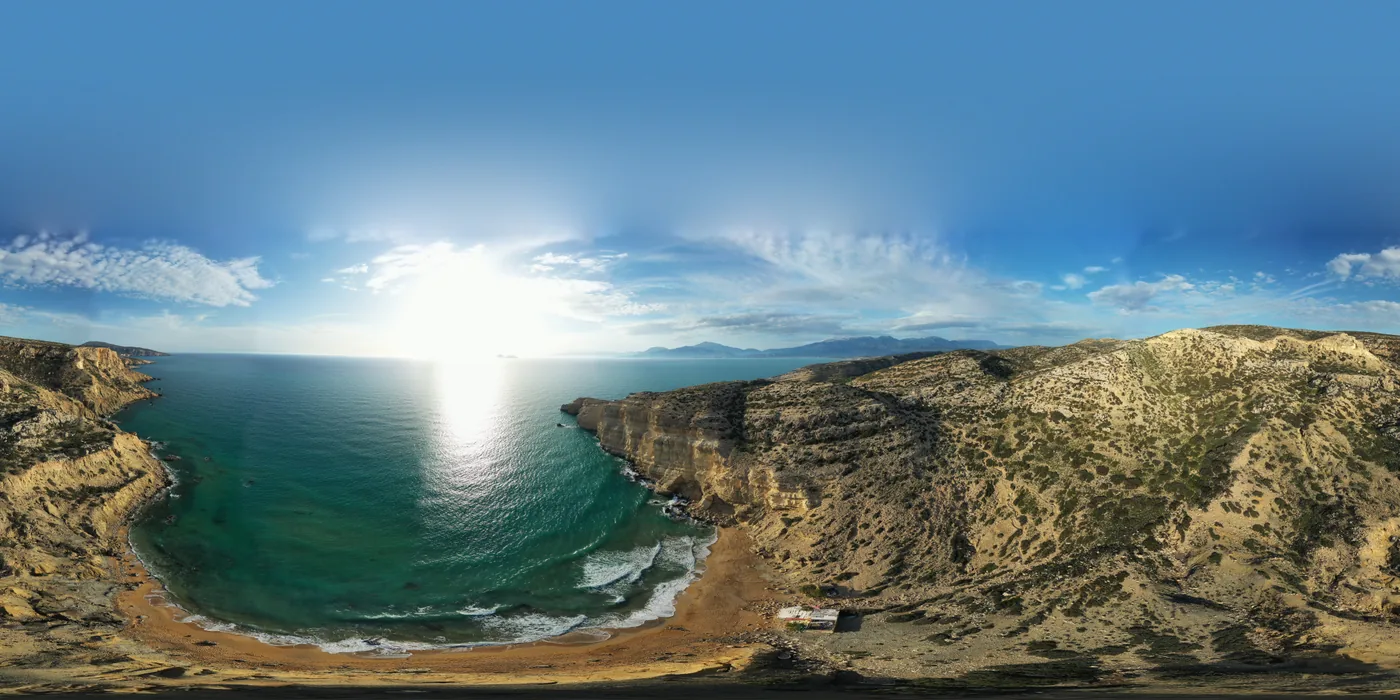 Aerial view of a small cove below sandstone cliffs, with a strip of reddish sand and the sun low over the sea.
