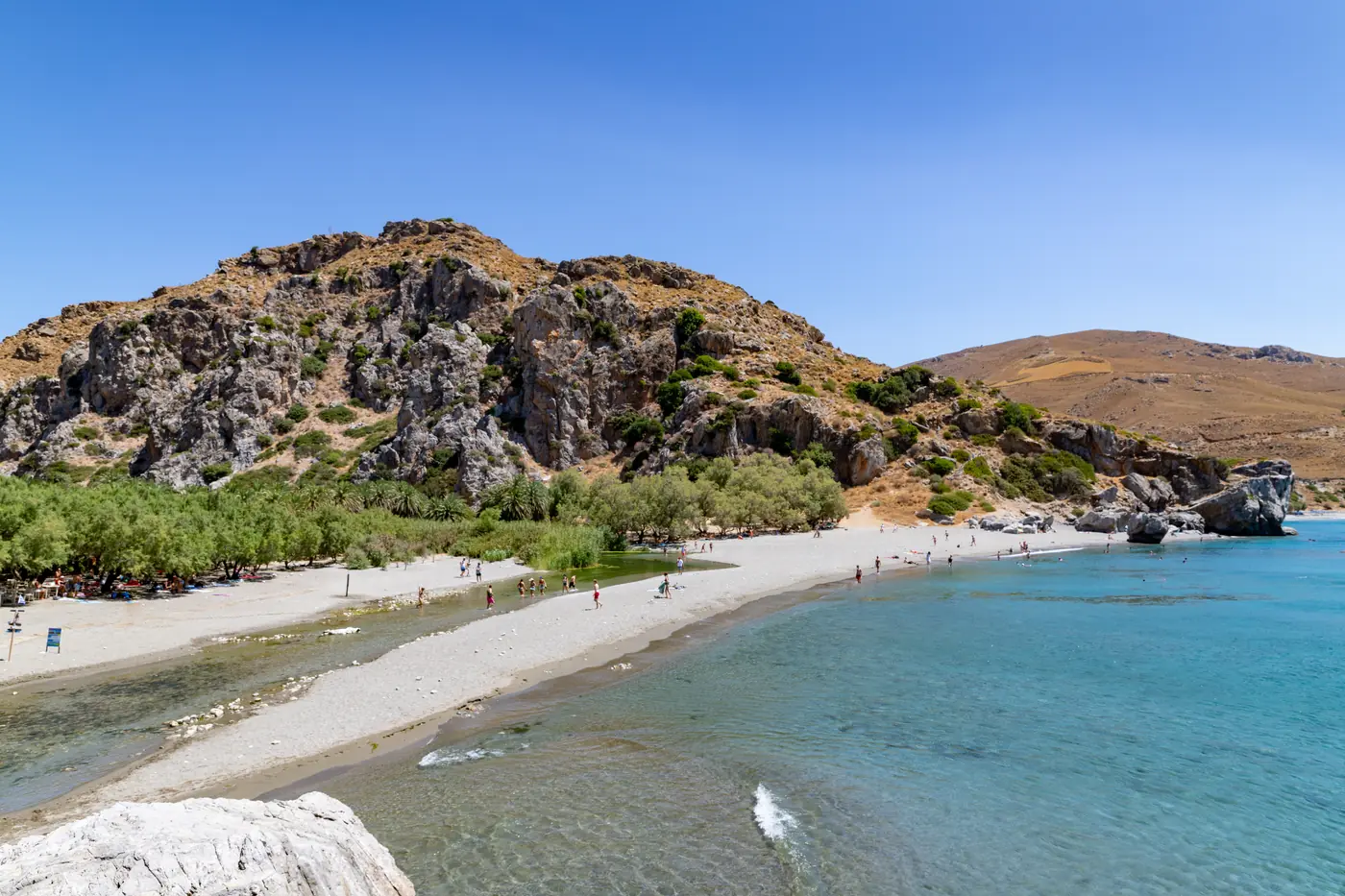 Aerial view of Preveli beach: a sand spit at a river mouth lined with palms, below steep rocky hills.
