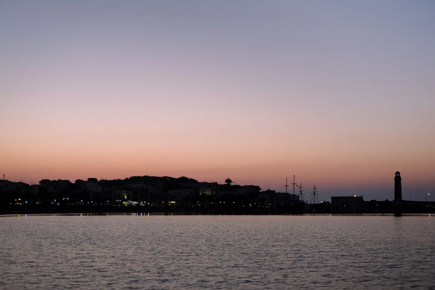 The old Venetian harbour of Rethymno and its lighthouse in silhouette against a pink evening sky over still water.