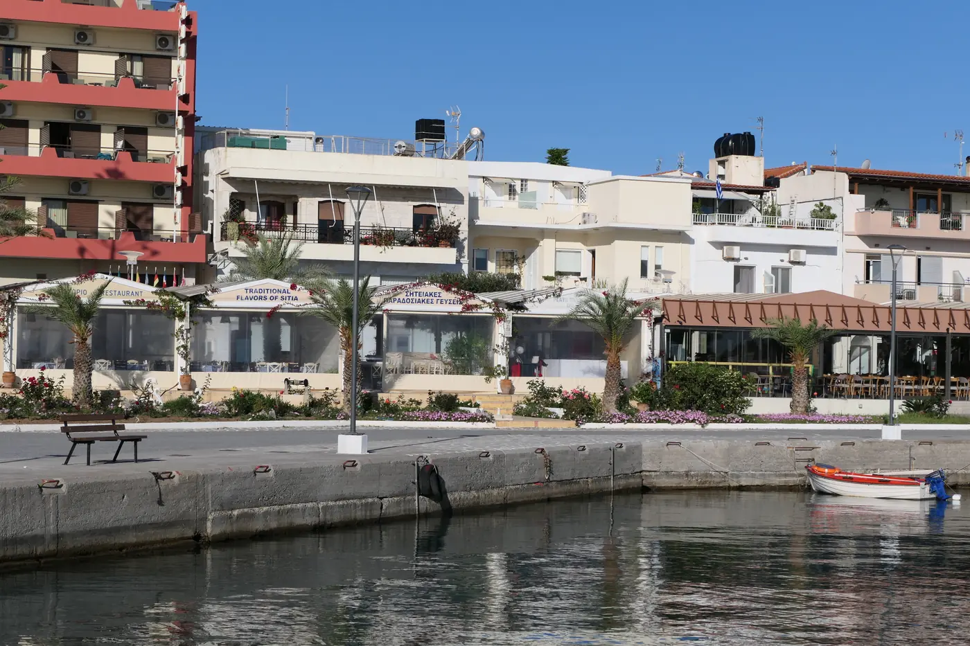 Sitia’s harbour front: a quay with a small boat moored, and a row of white apartment buildings with cafés and palm trees behind.
